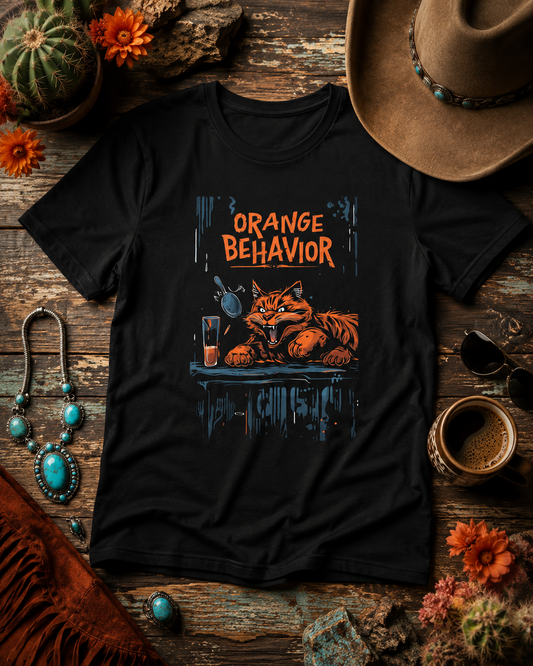 Black Orange Behavior cat graphic t-shirt styled on rustic wood with Southwestern desert accessories, cactus, turquoise jewelry, coffee, and a cowboy hat.
