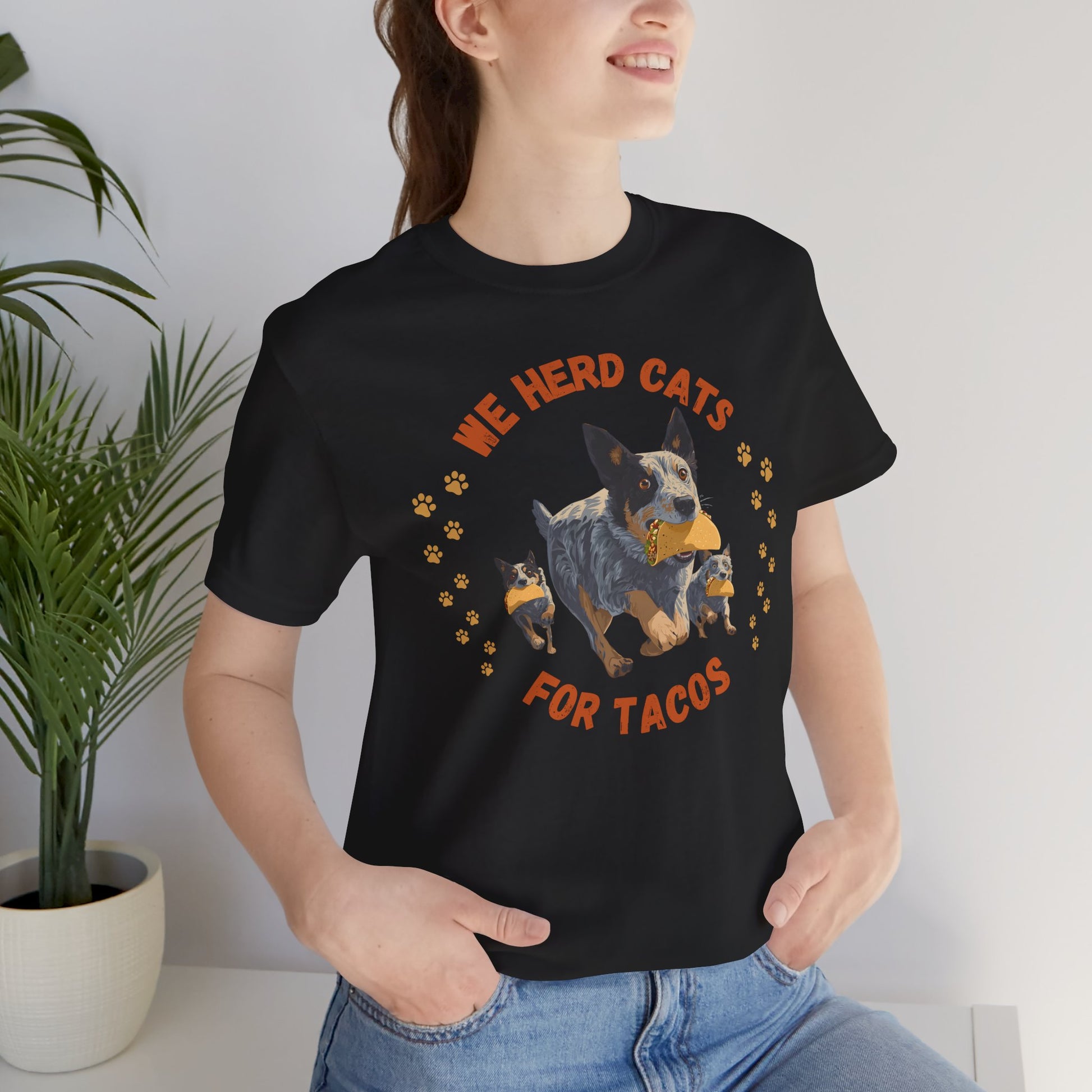 Person wearing a black t-shirt with Blue Heeler dog herding cats with tacos and text reading We Herd Cats for Tacos