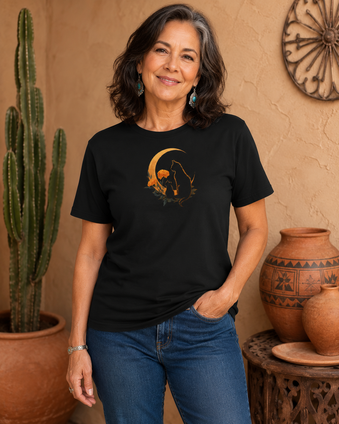 A woman wearing a black short-sleeve graphic t-shirt design with two silhouetted cats and a crescent moon, paired with blue jeans, standing in a rustic desert-themed room with a cactus and pottery.