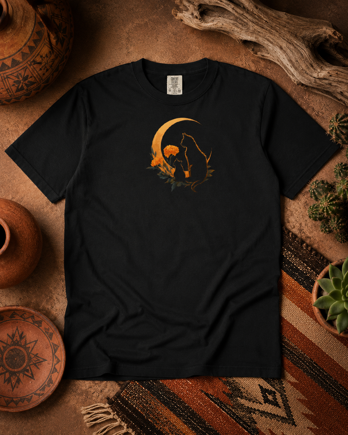 A black short-sleeve t-shirt with a graphic design with two silhouetted cats and a crescent moon and flowers, displayed in a rustic flat-lay setting with pottery, driftwood, and a patterned rug.