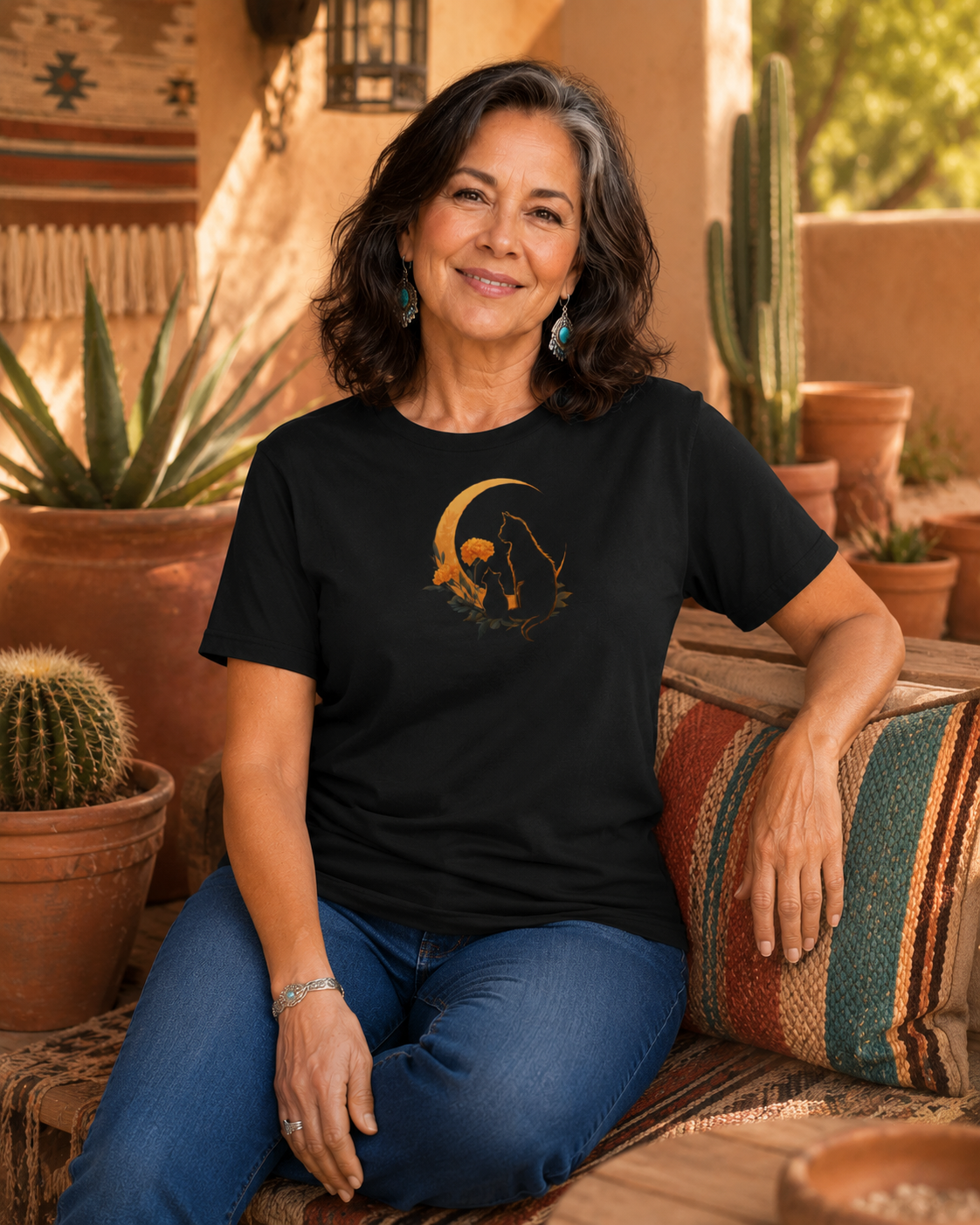 A woman sitting on a patterned couch wearing a black t-shirt design with two silhouetted cats and a crescent moon, set against a warm desert patio background with cacti.