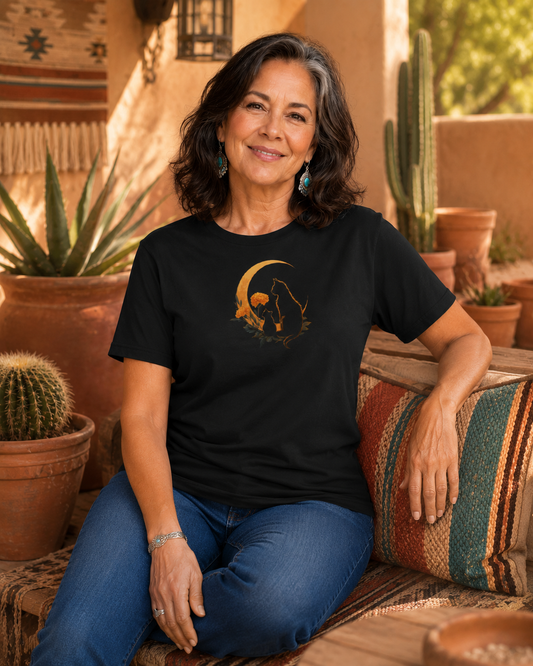 A woman sitting on a patterned couch wearing a black t-shirt design with two silhouetted cats and a crescent moon, set against a warm desert patio background with cacti.
