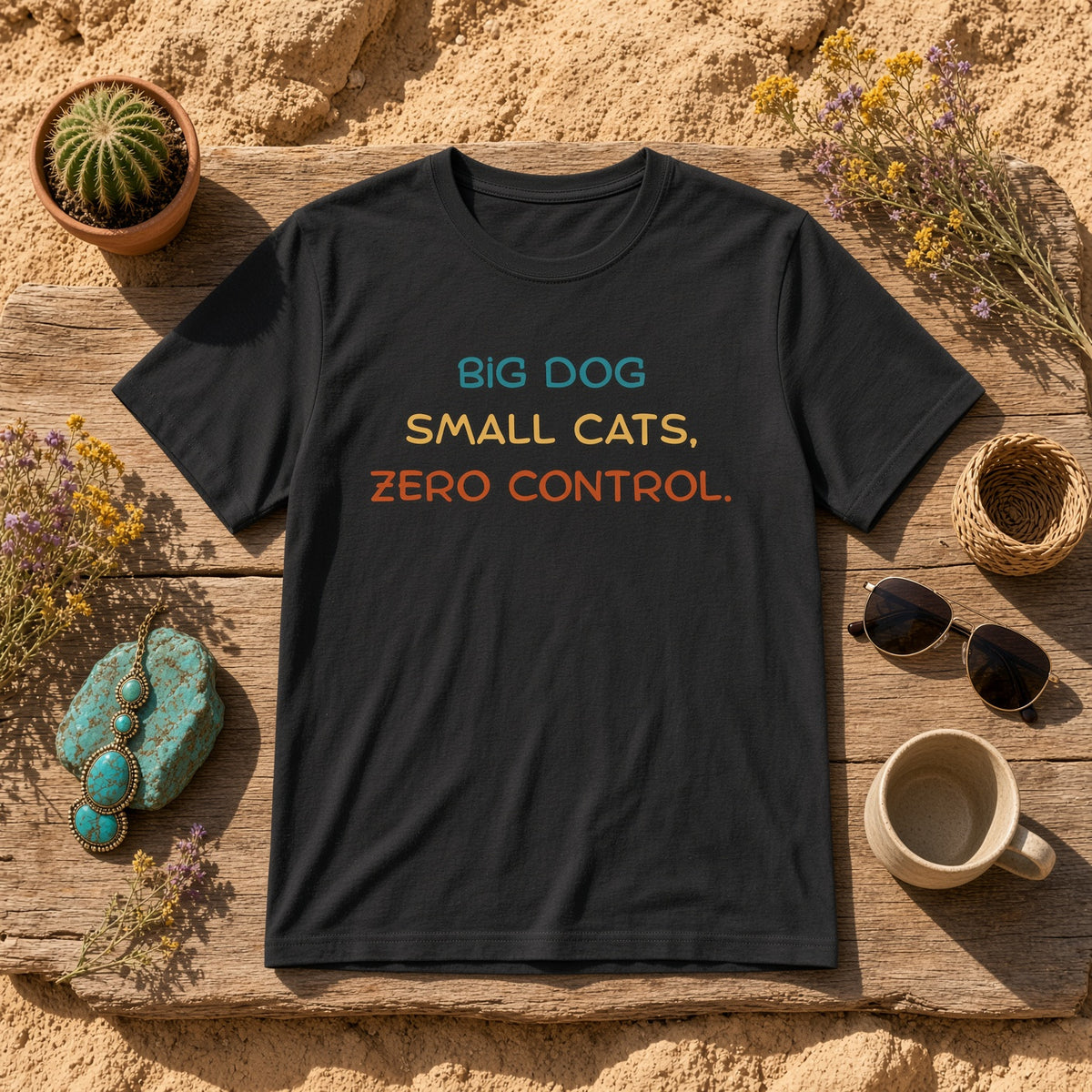 Black t-shirt on a wooden surface with text: 'BIG DOG SMALL CATS, ZERO CONTROL.' surrounded by decorative items.