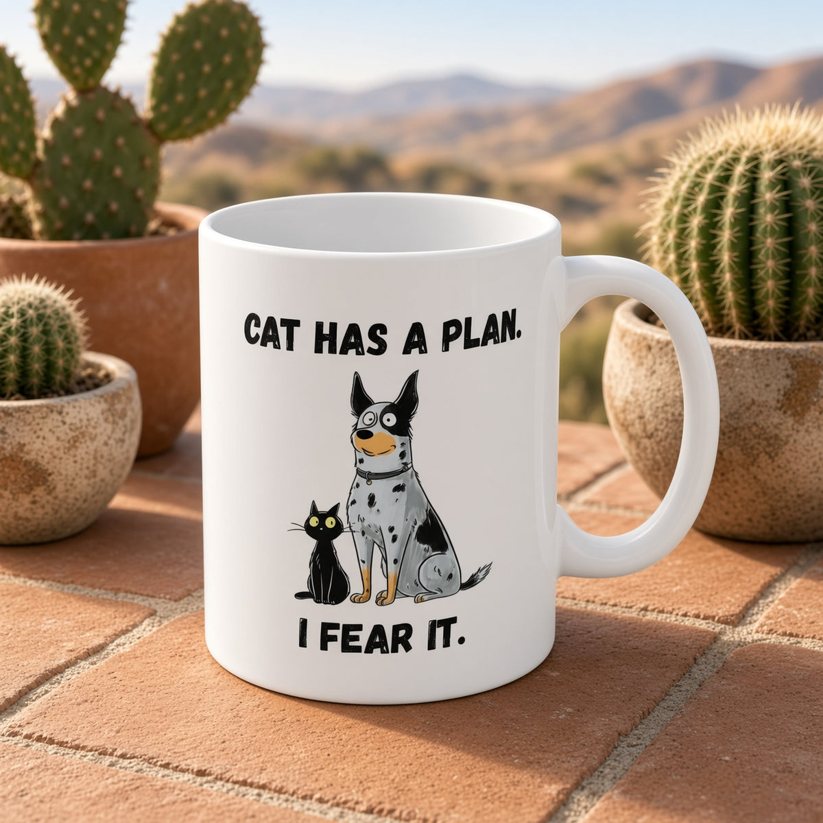 White mug featuring a dog and cat with 'CAT HAS A PLAN. I FEAR IT.' text, placed on a tiled surface with cacti.