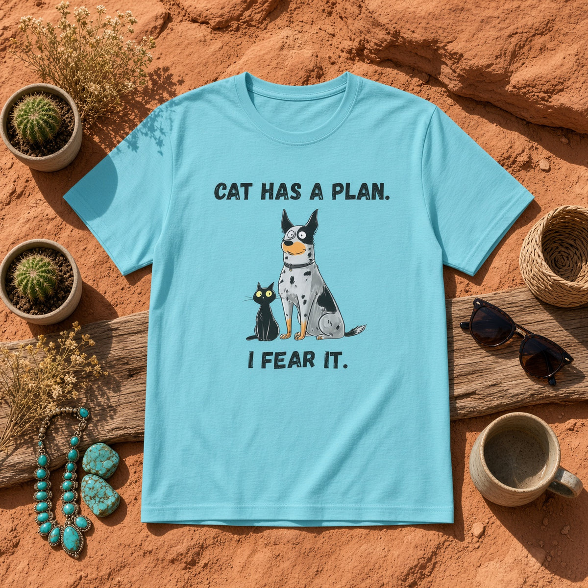 Flat lay of a blue t-shirt with a blue heeler and black cat design on a rustic background, text reads 'CAT HAS A PLAN. I FEAR IT.'
