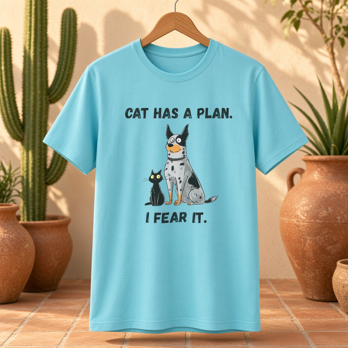 Blue t-shirt with a blue heeler and black cat design hanging next to cacti, text reads 'CAT HAS A PLAN. I FEAR IT.'