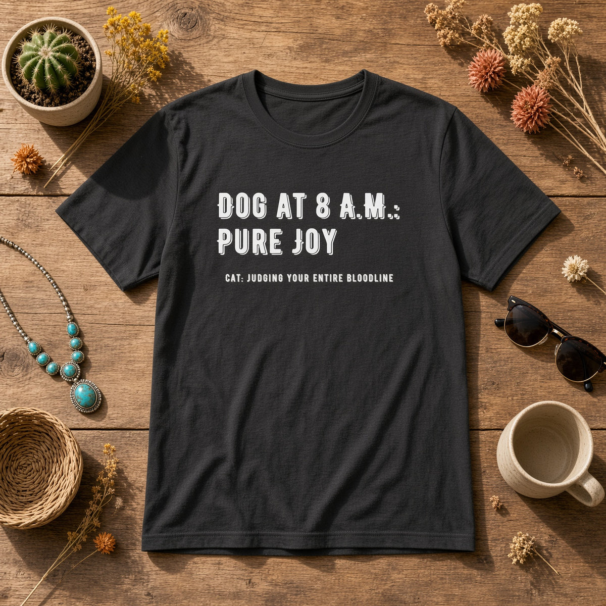 Black t-shirt laid flat on a wooden surface with the text 'DOG AT 8 A.M.: PURE JOY' and 'CAT: JUDGING YOUR ENTIRE BLOODLINE', surrounded by decorative items