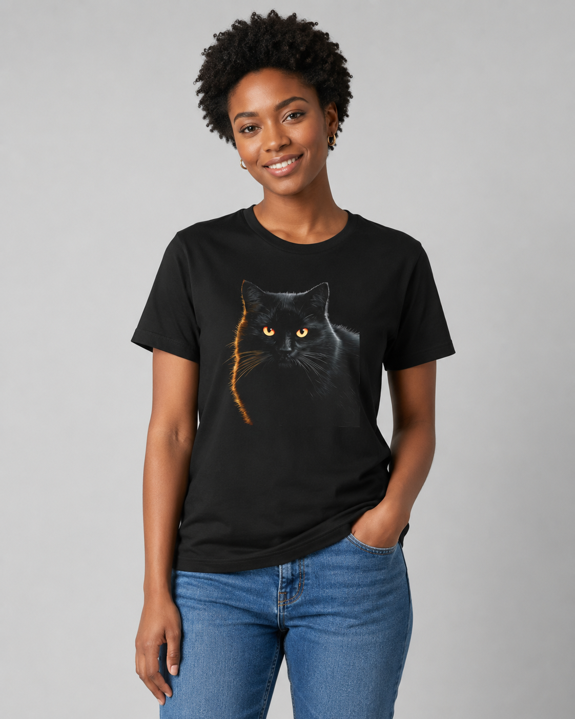A woman wearing a black short-sleeve crewneck t-shirt with a graphic of a black cat with glowing yellow eyes and a gold tail, paired with blue jeans, against a solid gray studio background.
