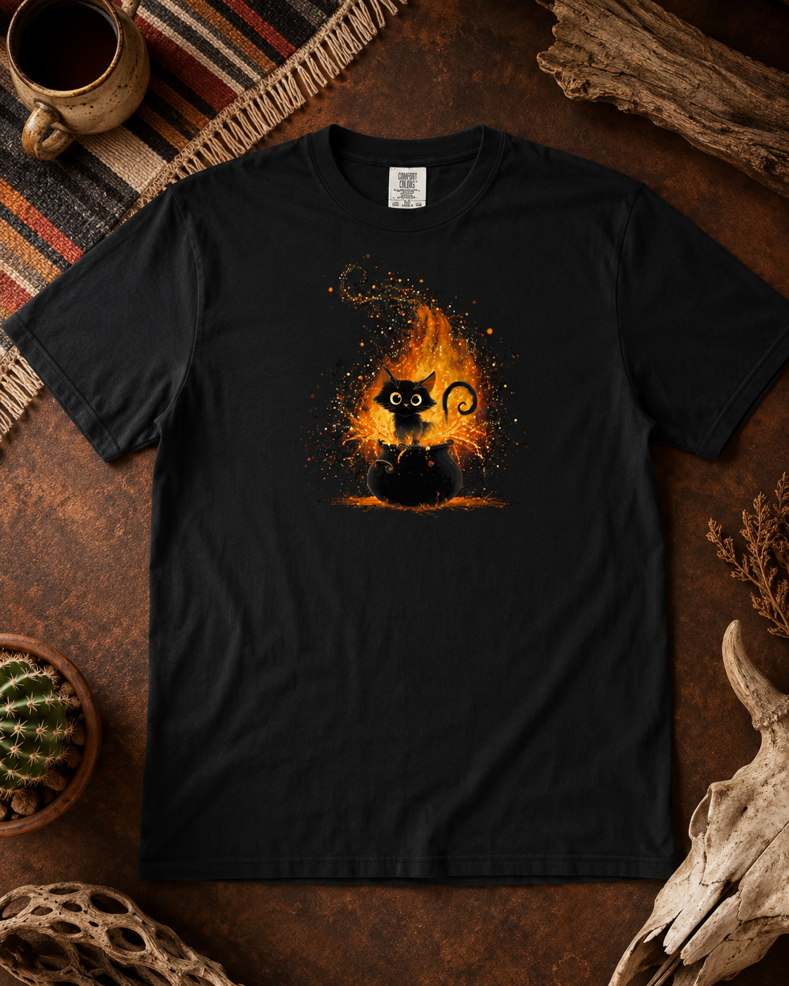 A black short-sleeve t-shirt laid flat on a rustic brown surface, featuring a graphic of a black cat surrounded by glowing orange and yellow flames. The shirt is styled with a mug, woven mat, dried plant, and small cactus in a cozy flat-lay arrangement.