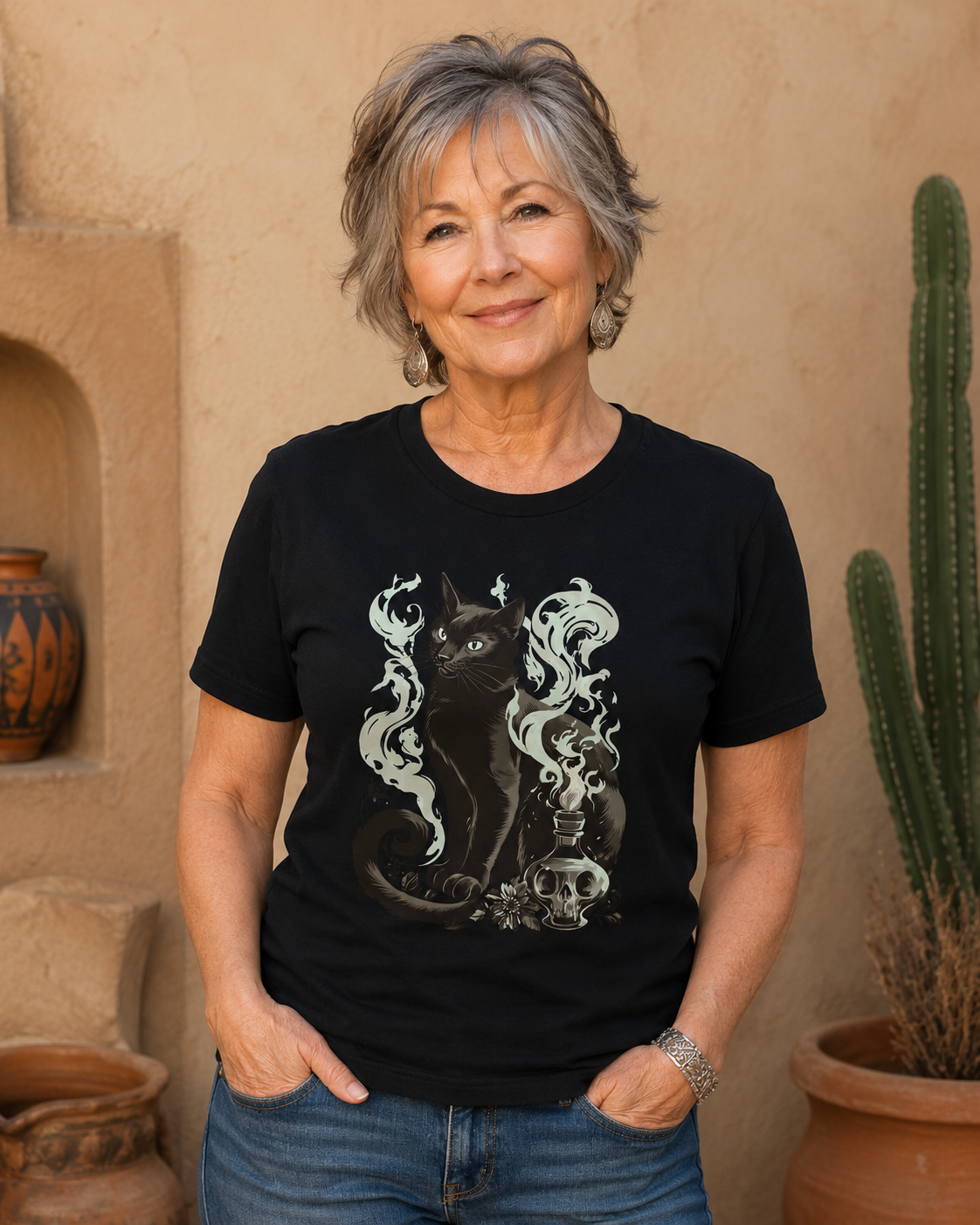 A woman with short gray hair wearing a black graphic t-shirt featuring a black cat and white smoke design, standing in front of a beige adobe wall with a cactus in a pot.