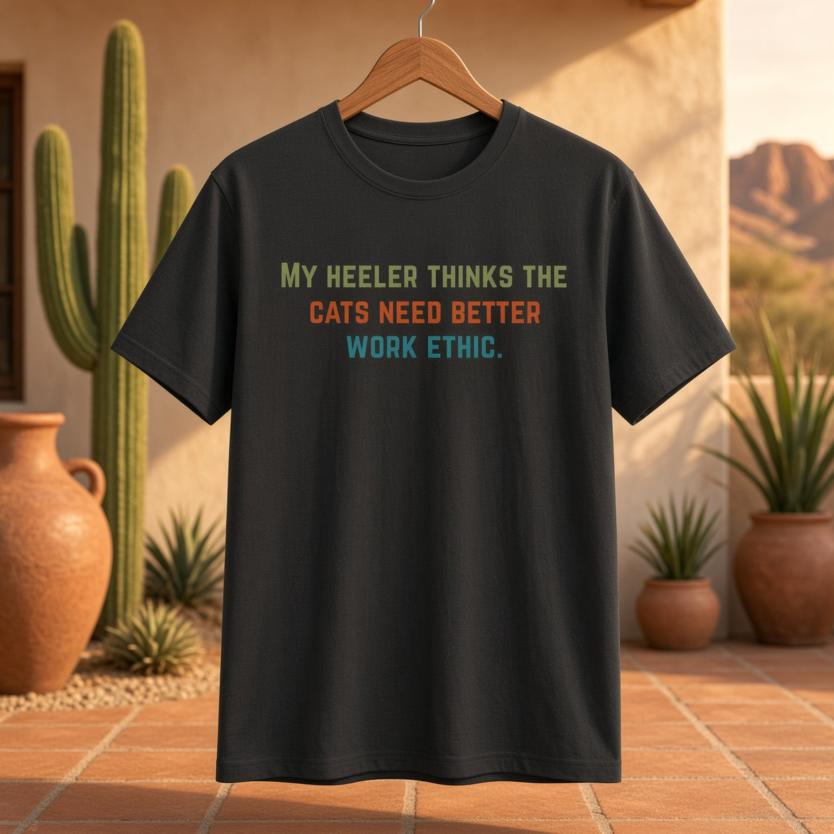 Black t-shirt hanging on a wooden hanger in a desert-themed setting with the text 'My heeler thinks the cats need better work ethic.'