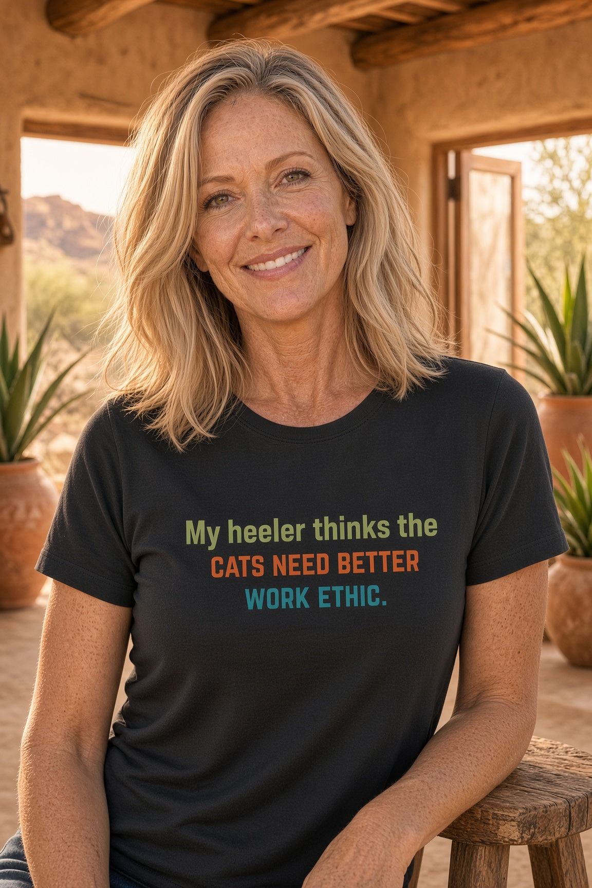 Woman smiling in a black t-shirt with the text 'My heeler thinks the cats need better work ethic.'
