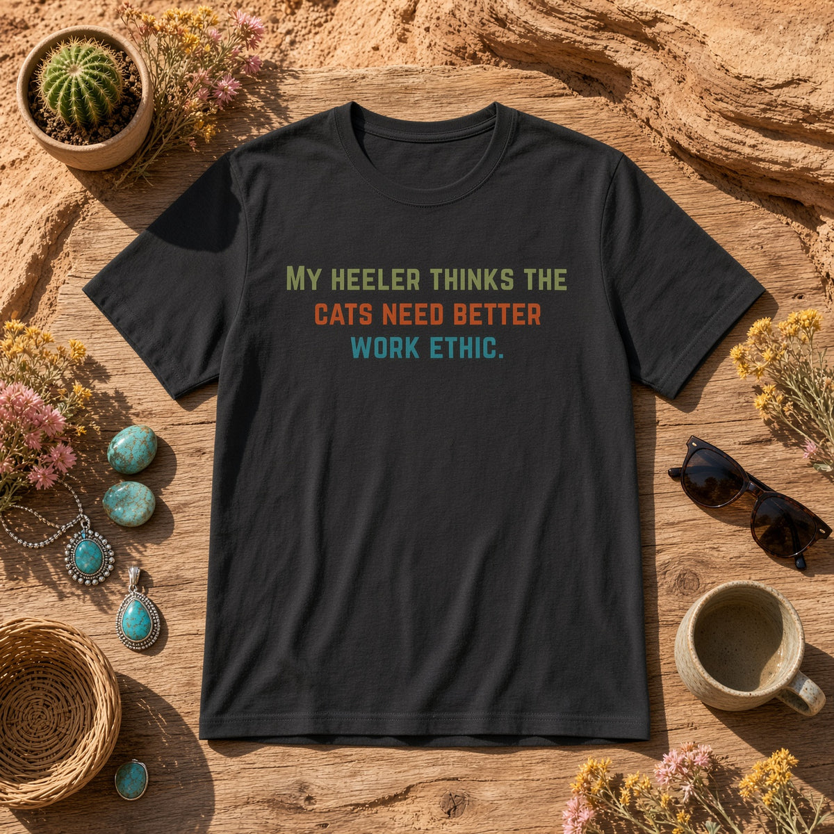 Black t-shirt laid with the text 'My heeler thinks the cats need better work ethic.' laid flat on a rustic surface.