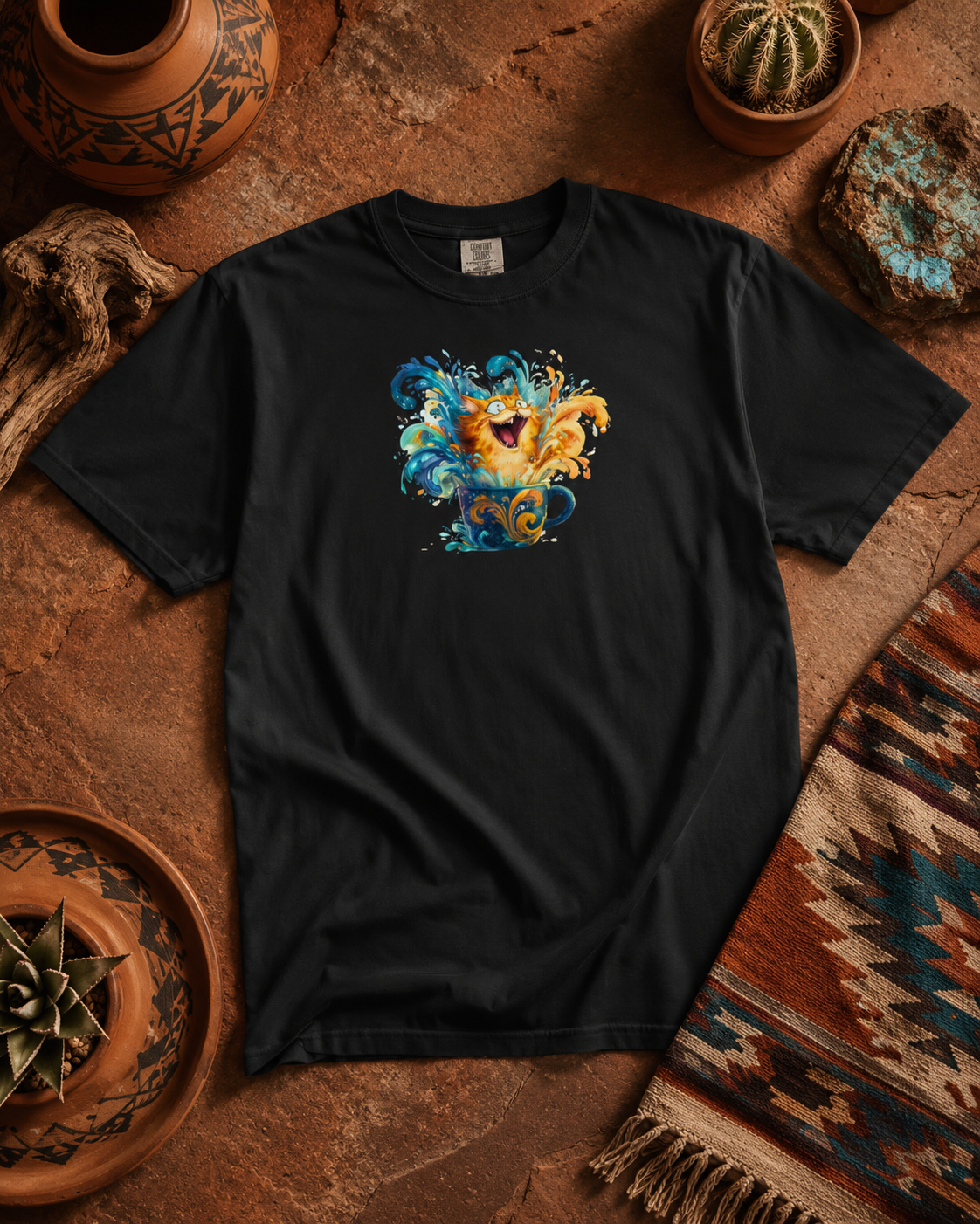 A black short-sleeve t-shirt with a colorful graphic of a ginger tabby bursting through a spray of bright blue and orange chaos in a mug., displayed in a styled flat-lay on a rustic brown stone surface surrounded by pottery, cacti, and a patterned textile.
