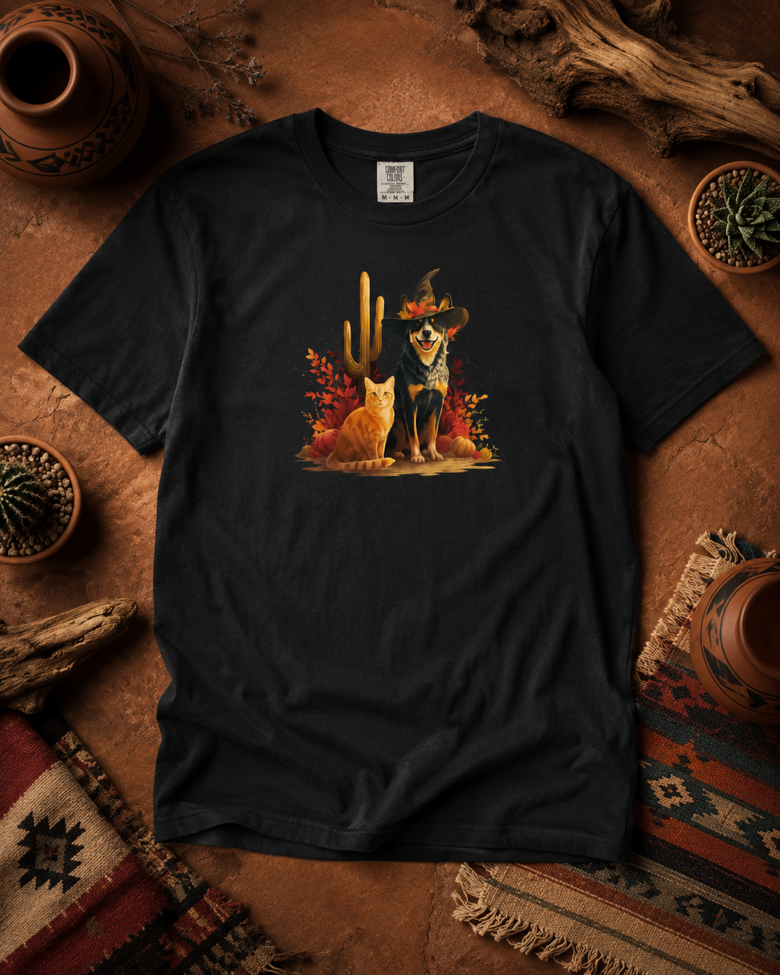 A black short-sleeve t-shirt laid flat on a rustic brown surface, featuring a colorful graphic of a orange cat and heeler in a witch hat desert autumn feels The shirt is styled with pottery, dried wood, and patterned textiles around it.