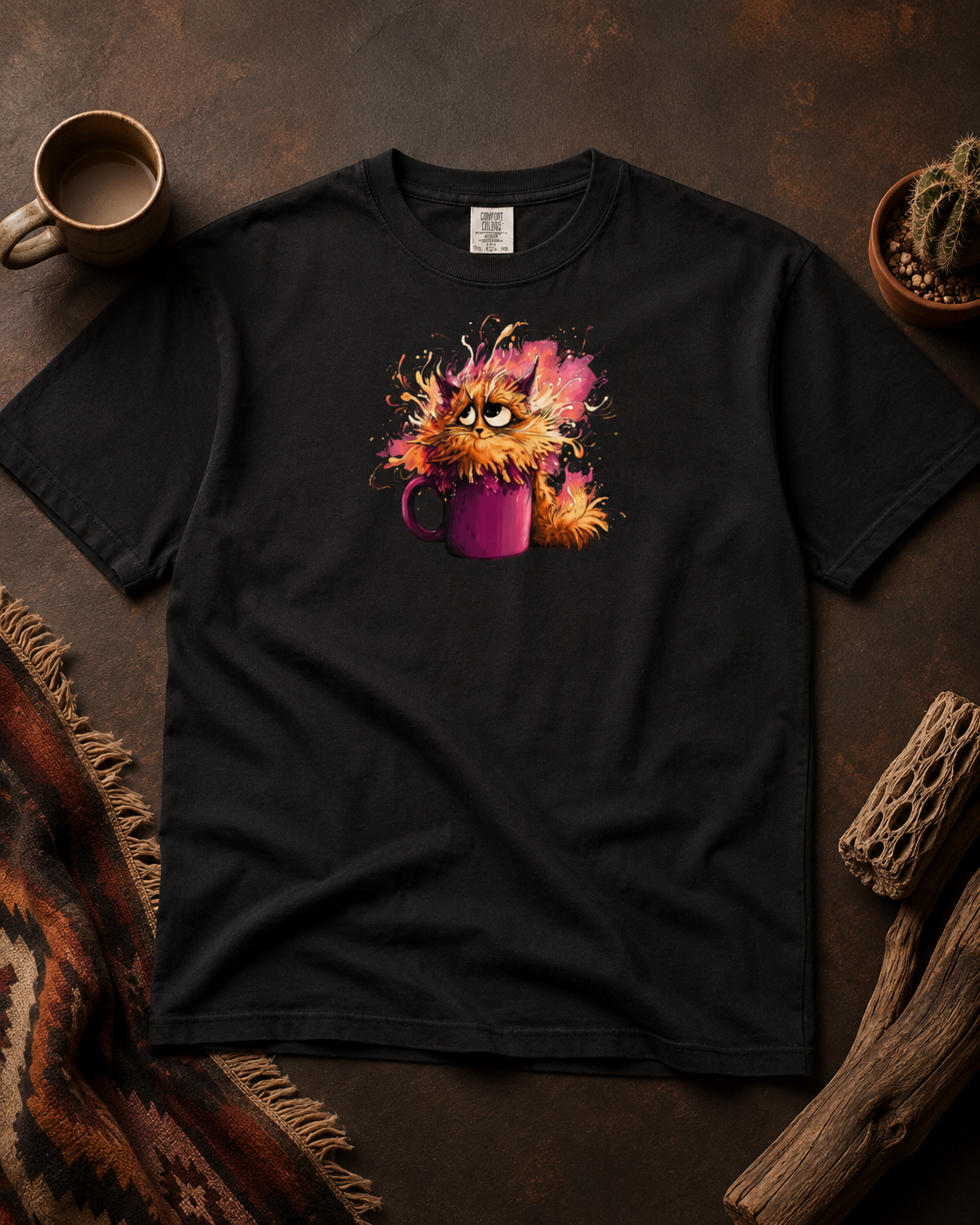 A black short-sleeve t-shirt laid flat on a rustic brown surface, featuring a colorful graphic of a fluffy orange cat sitting in a purple mug with pink and yellow paint-like splatters. A coffee cup, plant, and wood decor are arranged around it.