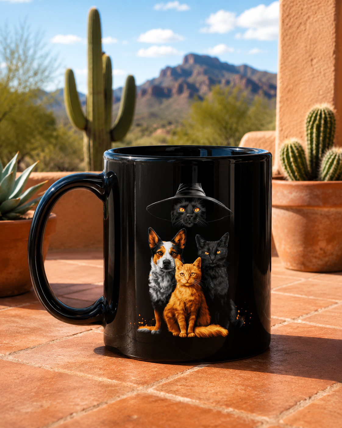 A black ceramic mug with a printed Black mug with a graphic of a black cat in a witch hat with a cattle dog, a black cat  and a orange cat., displayed on a terracotta patio tile with desert plants and mountains in the background.