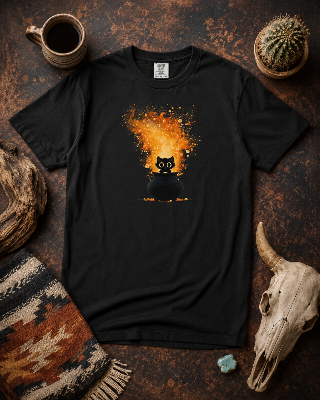 A black short-sleeve t-shirt laid flat on a rustic dark brown surface, featuring a graphic of a black cat sitting in a caldron of a bright orange and yellow fire. The shirt is styled with a mug, cactus, woven blanket, skull, and stones.
