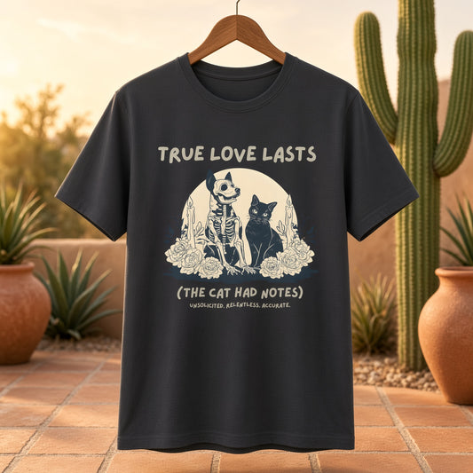 Black tee with a skeleton dog and black cat design with the text 'True Love Lasts (The Cat Had Notes)' hanging on a wooden hanger outdoors.