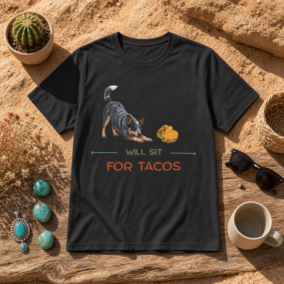 Black t-shirt featuring a Blue Heeler and taco graphic with the text 'WILL SIT FOR TACOS' on a sandy surface with decorative items.