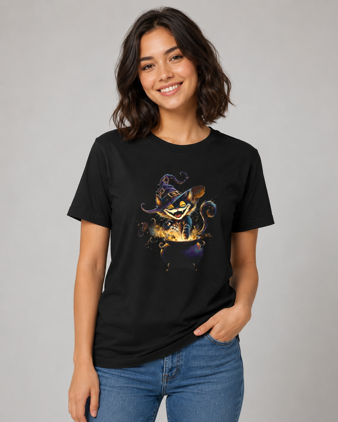 A woman wearing a black short-sleeve graphic t-shirt with a colorful illustration of a cat in a cauldron, paired with blue jeans, against a solid gray studio background.