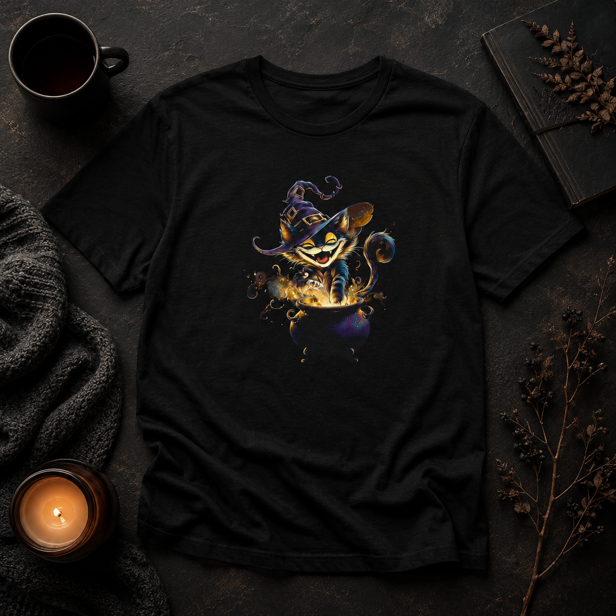 A black short-sleeve graphic T-shirt laid flat on a dark textured surface, surrounded by cozy props including a mug, candle, knit fabric, notebook, and dried flowers. The shirt features a colorful central illustration of a cat character in a witch hat emerging from a cauldron of chaos.