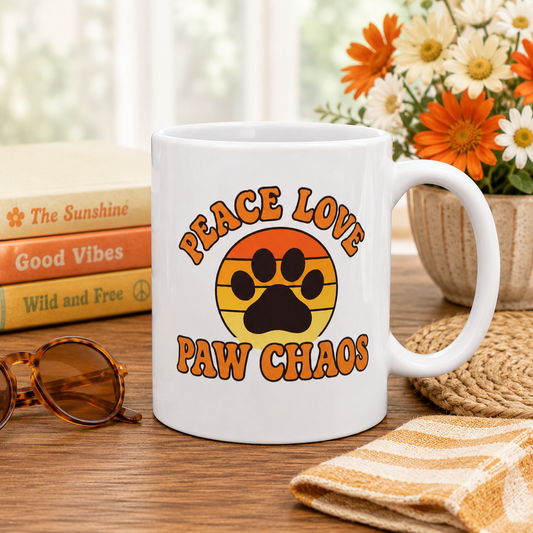 White mug with 'Peace Love Paw Chaos' design on a wooden surface with books and flowers in the background.