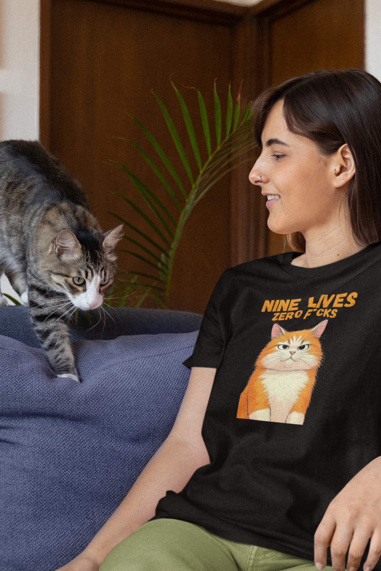 Woman wearing a black t-shirt with a cat graphic and text, sitting on a couch next to a cat.